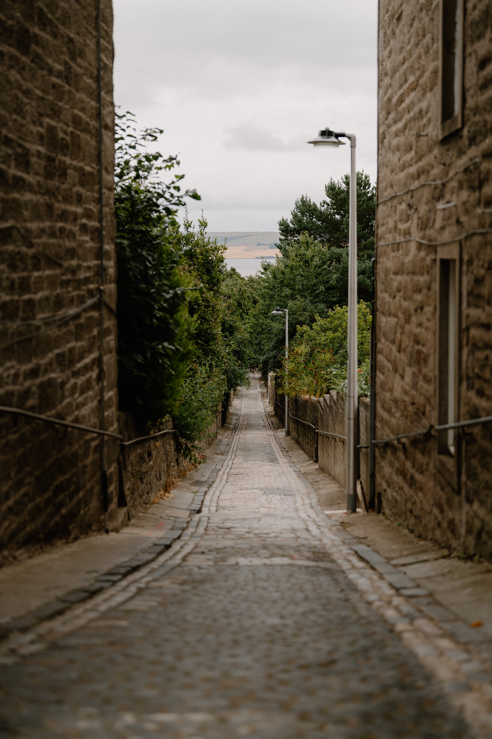 A narrow cobbled lane descends between stone buildings towards a distant firth, a lamp post standing partway down.