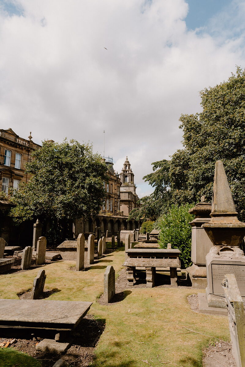 Table tombs and headstones fill a churchyard, a domed clock tower and cathedral spire visible above the trees beyond.