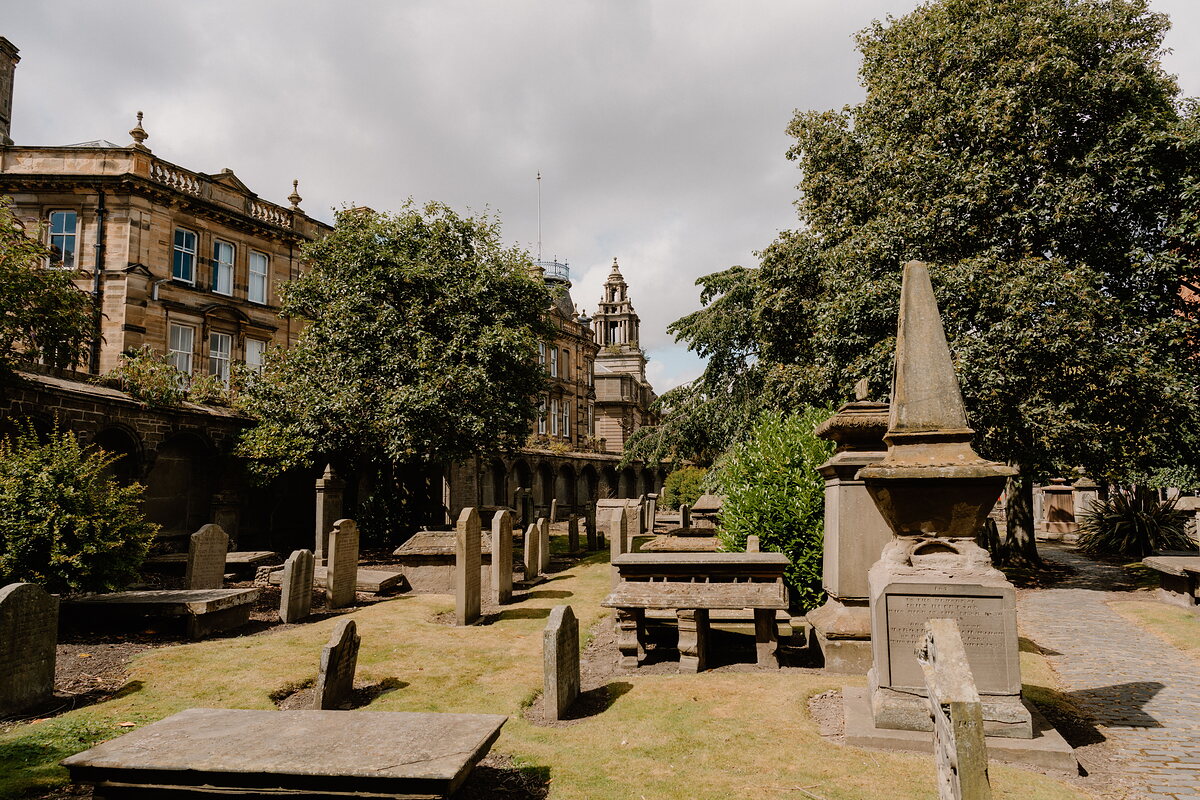 A stone obelisk stands among table tombs and headstones in a hillside cemetery, city buildings and a domed tower visible beyond the trees.