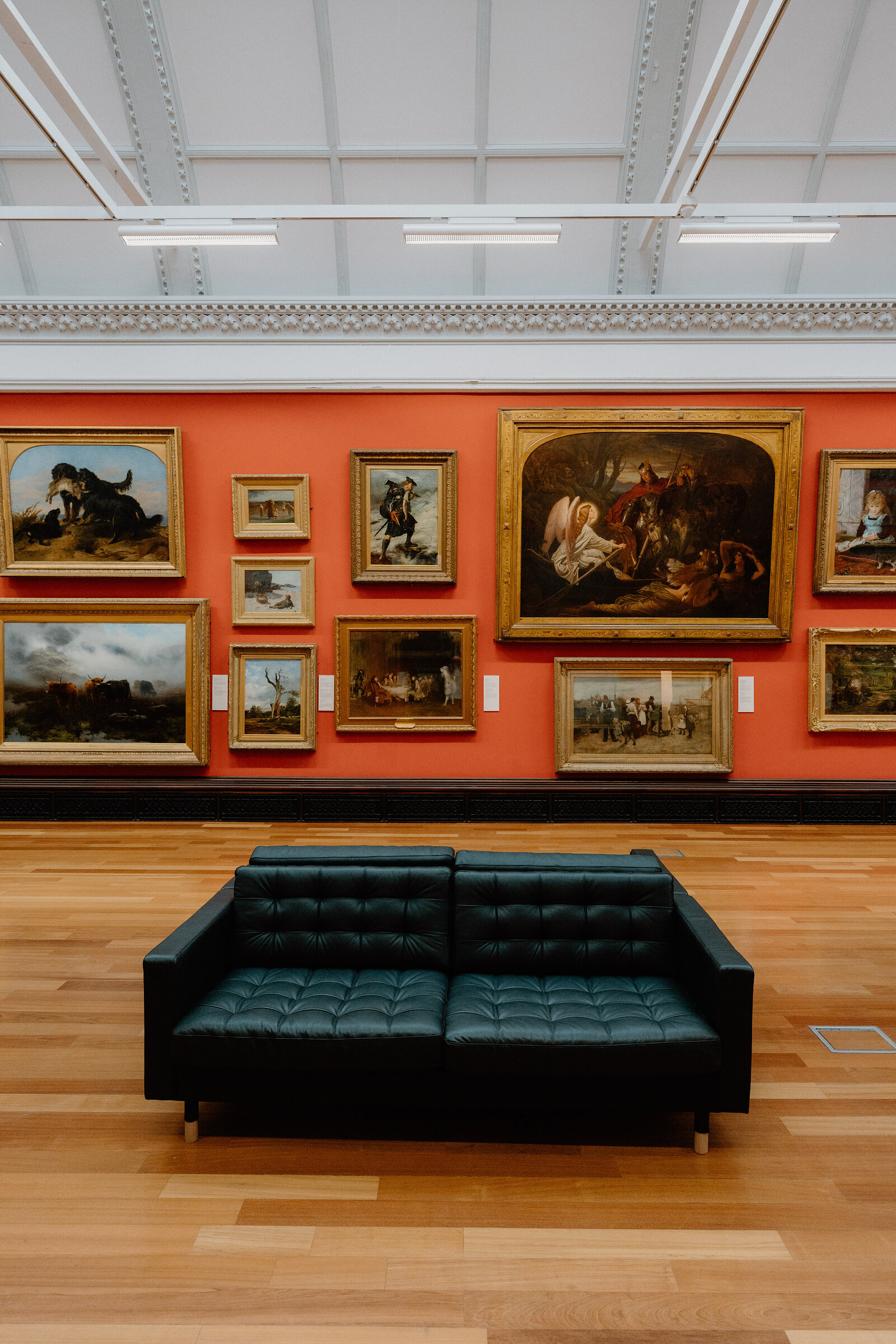 Gilt-framed oil paintings hang on a terracotta wall in a gallery, a black leather bench standing on the wooden floor in front.