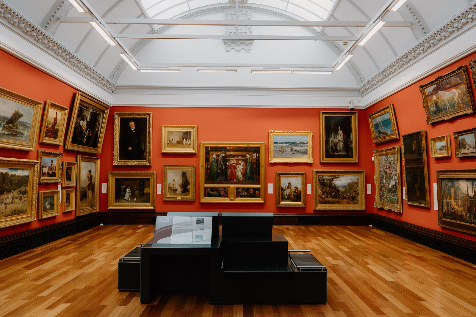 Gilt-framed oil paintings hang on a terracotta wall beneath a vaulted skylight in a gallery, an information desk standing on the wooden floor.