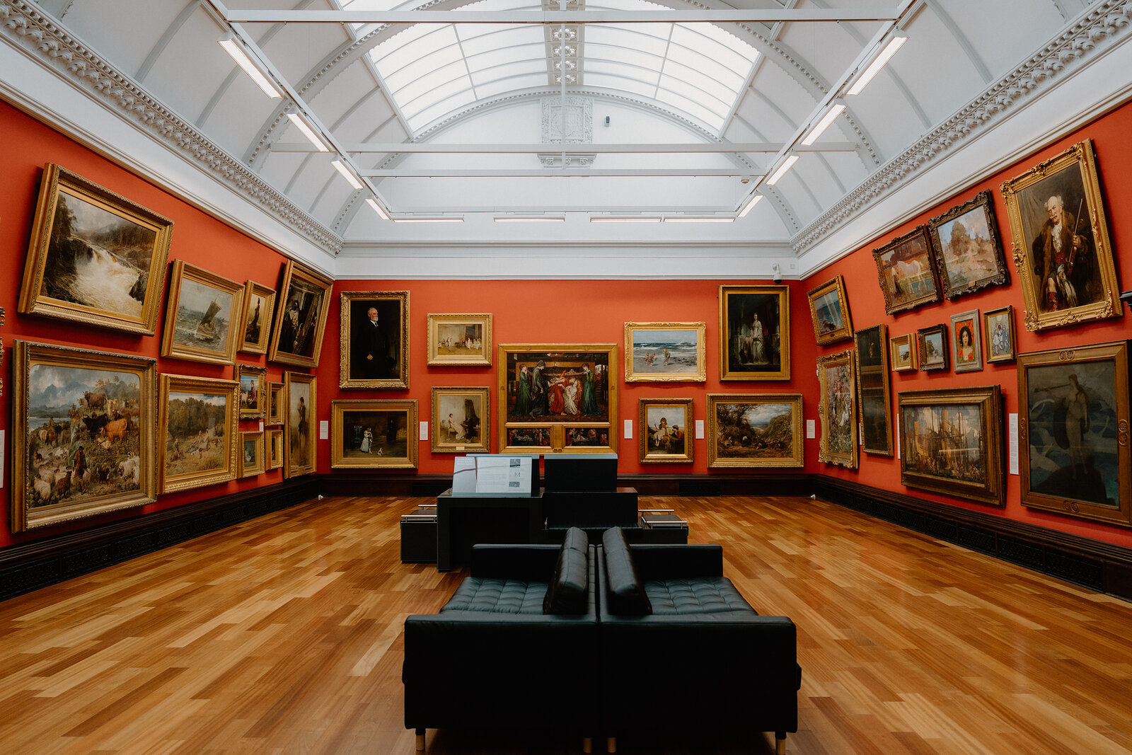 Gilt-framed oil paintings cover a terracotta wall in a gallery, black leather seating arranged in the centre of the wooden floor.