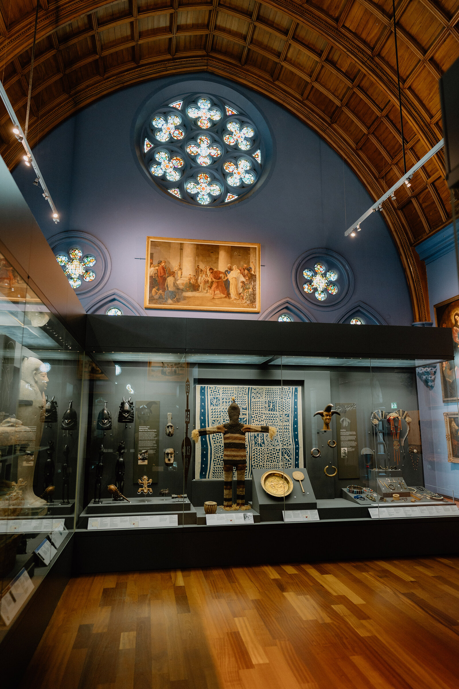 A rose window of stained glass glows above a museum display case holding a suit of chainmail, jewellery and textiles, a framed painting hanging between arched windows.