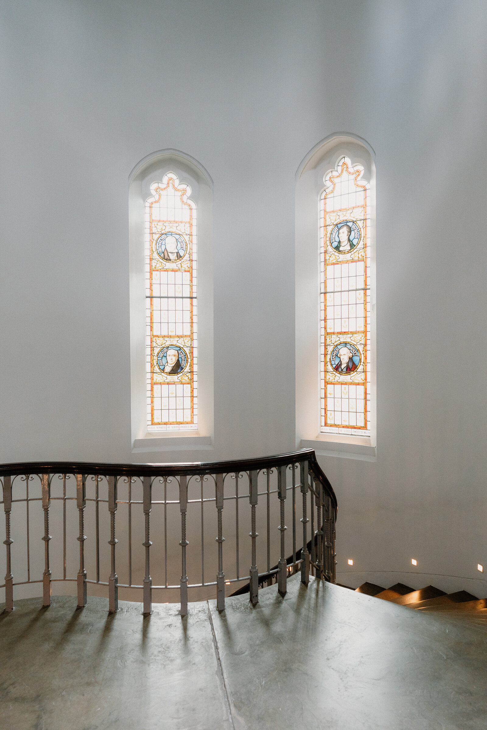Two tall stained glass windows with circular portrait medallions light a museum stairwell, an ornate iron railing curving above the stairs.