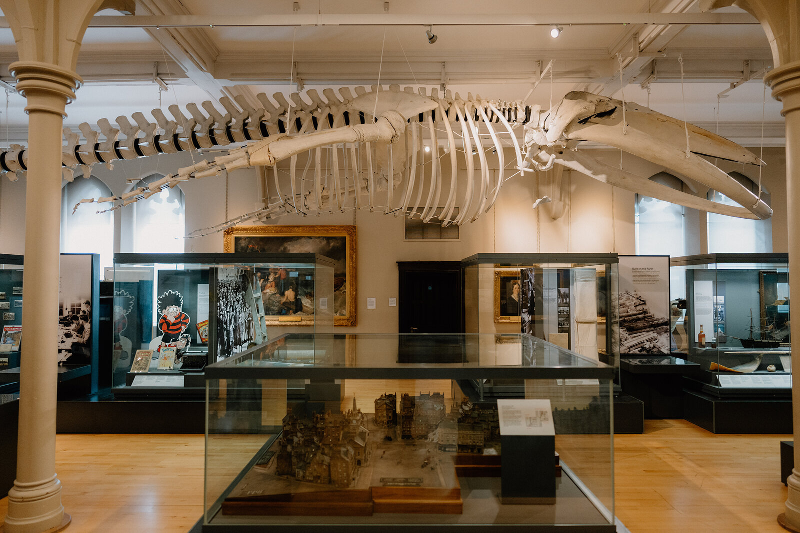 A whale skeleton hangs suspended above glass display cases in a museum hall, framed paintings and model ships arranged beneath it.