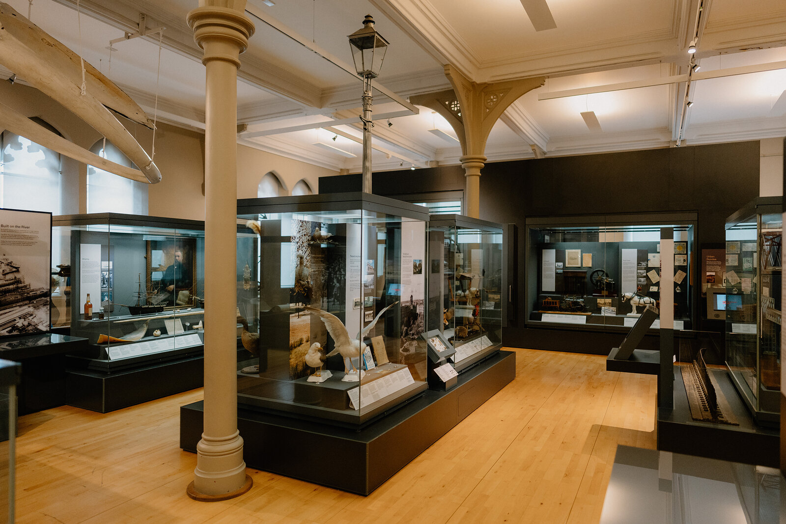 Glass display cases stand beneath an ornate cast-iron column in a museum hall, a hanging lantern and vaulted ceiling visible above.