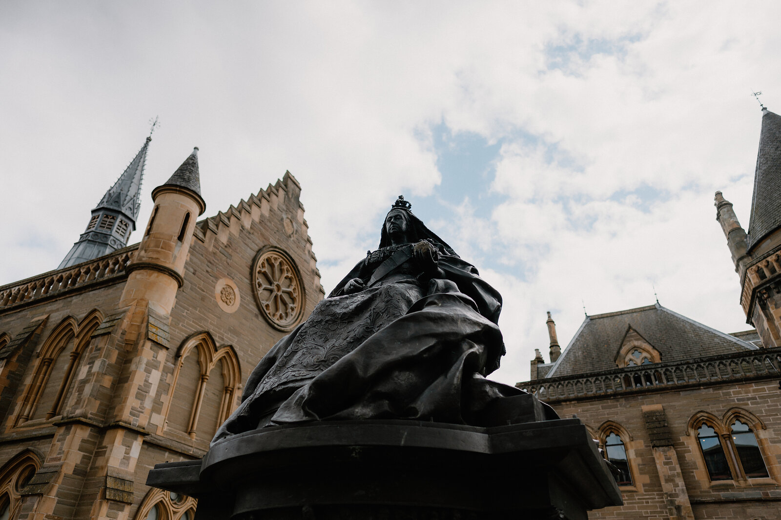 A dark bronze statue of a seated queen stands before a gothic sandstone building with a turreted tower and rose window.