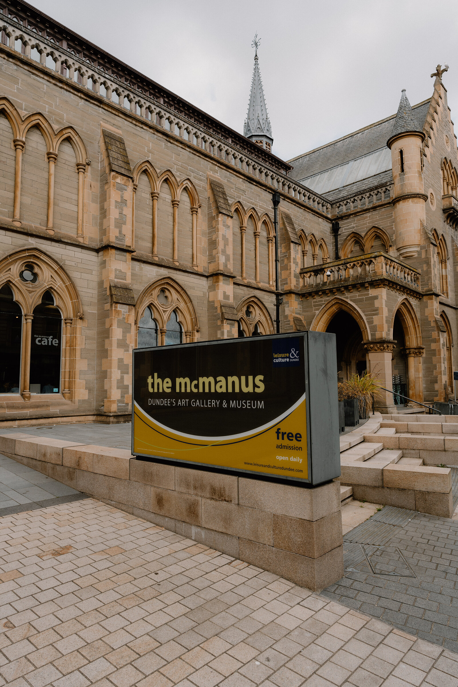 A tall gothic sandstone building with turrets and pointed arch windows, a café at ground level, and a sign in the foreground reading 'the mcmanus, Dundee's Art Gallery & Museum, free'.