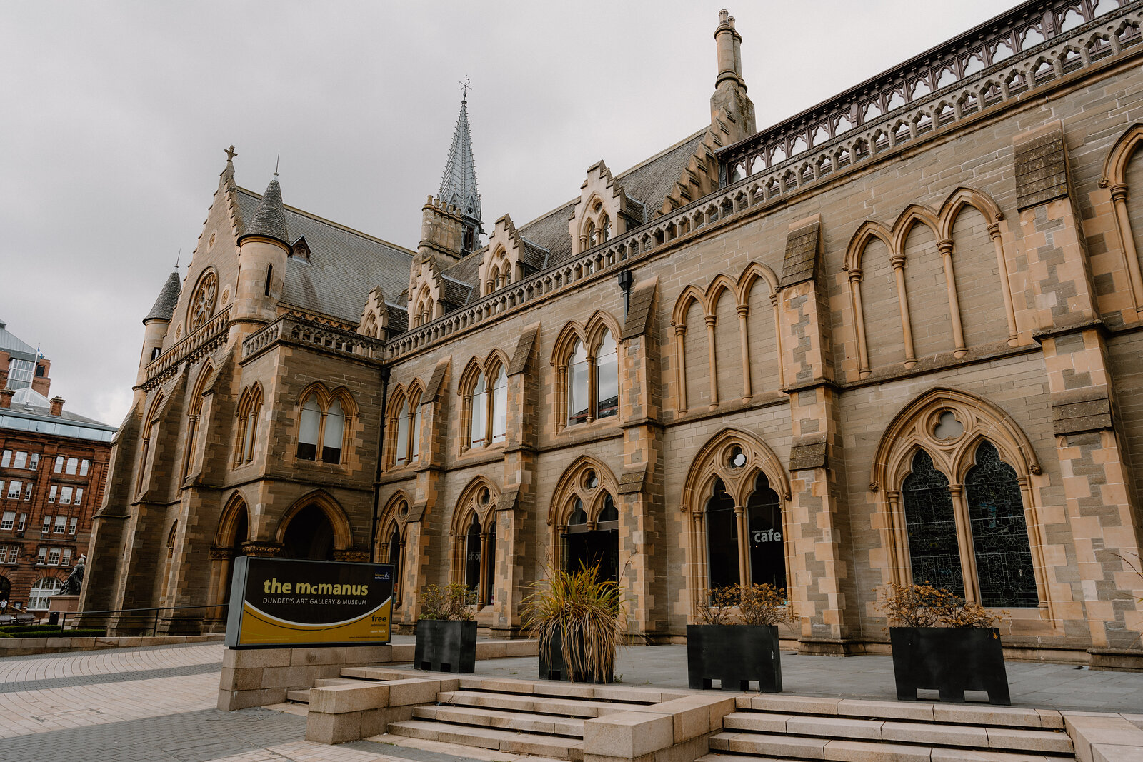 A gothic sandstone building with turrets, pinnacles and pointed arch windows rises above a paved plaza with a sign reading 'the mcmanus' beside a café entrance.