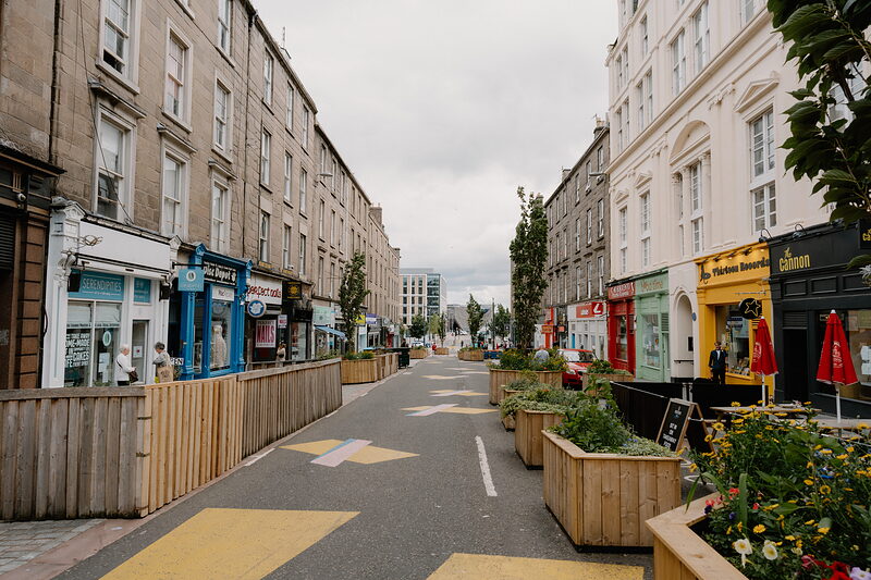 A pedestrianised street with painted road markings and timber planters runs between tenement buildings and shopfronts under an overcast sky.
