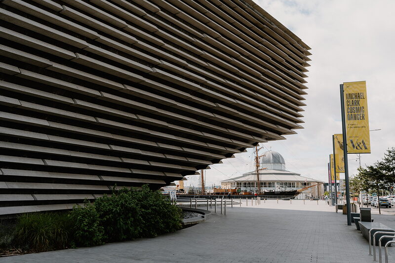 The tiered, ribbed concrete walls of a modern museum building curve toward a domed glasshouse and a ship's mast visible along the waterfront.
