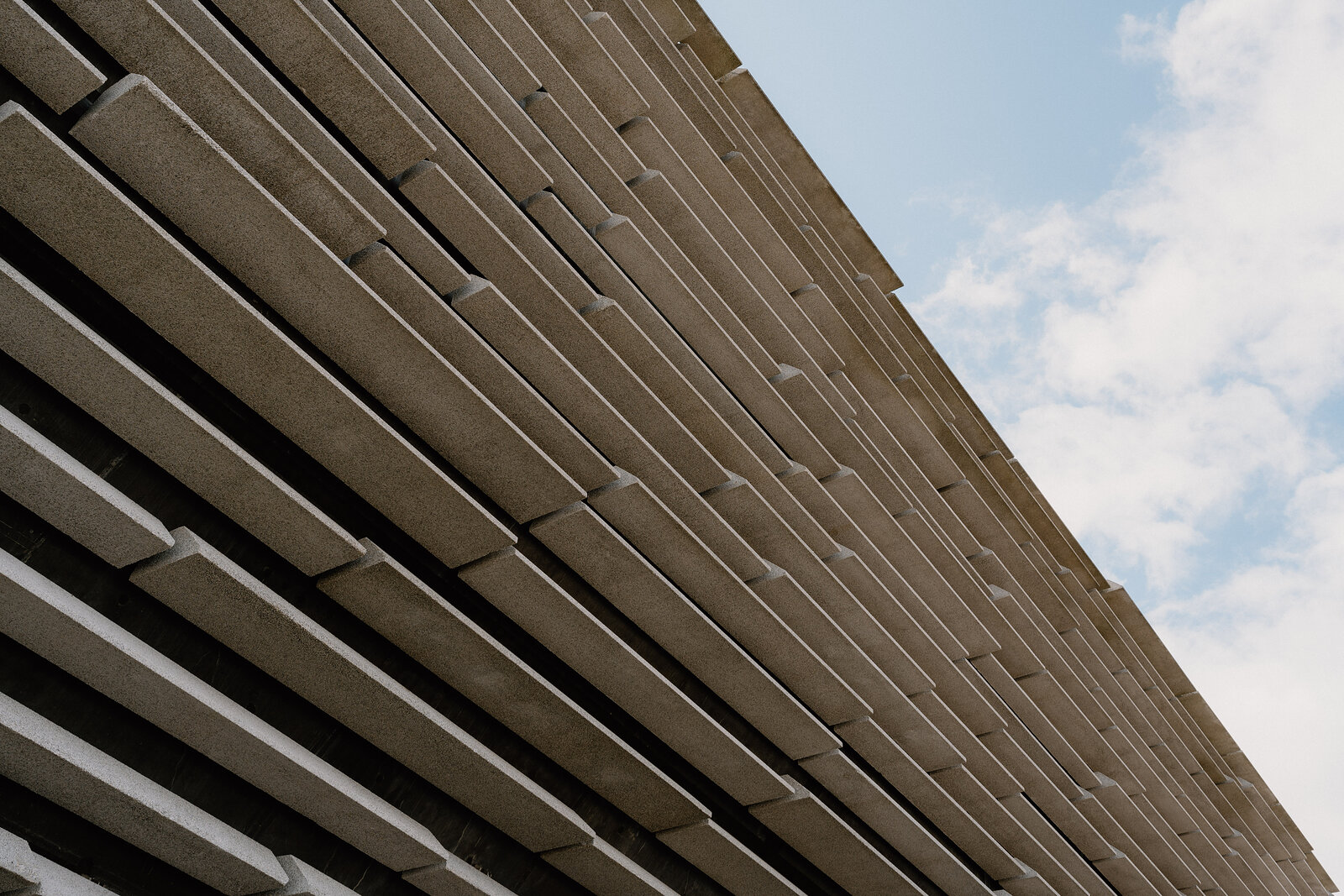 Looking up at a modern building's tiered concrete cladding as it recedes in ribbed rows toward a pale sky with scattered cloud.