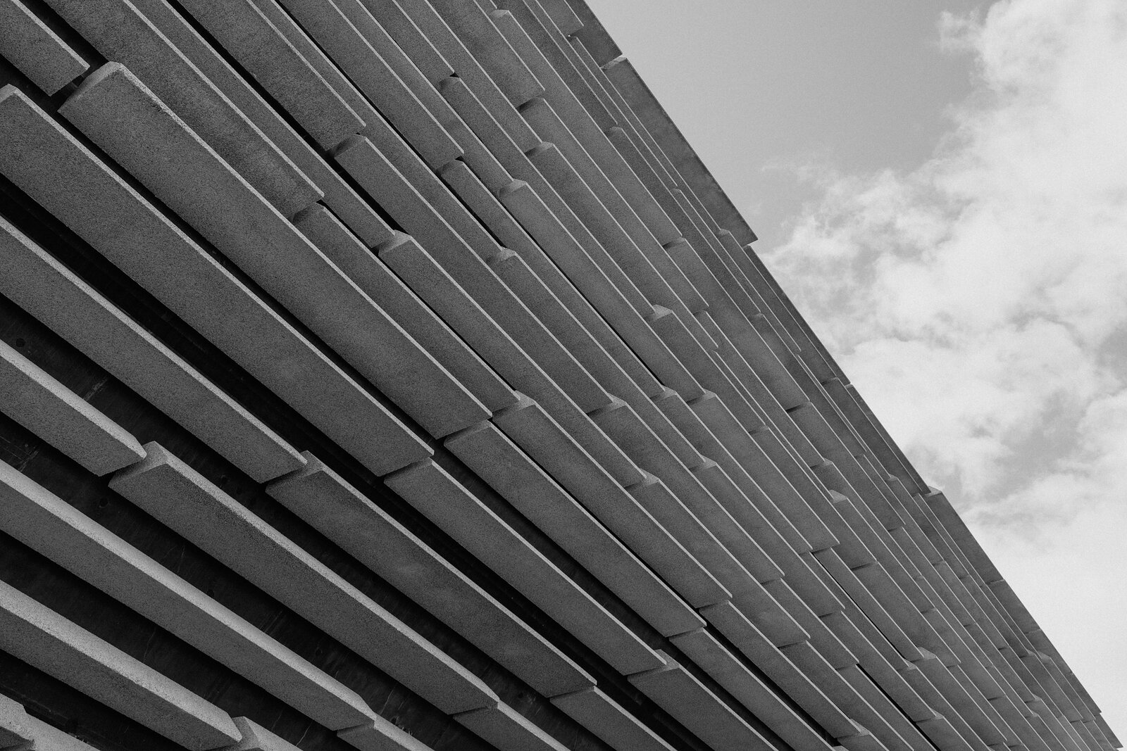 A monochrome view looking up at a modern building's tiered concrete cladding against a partly clouded sky.