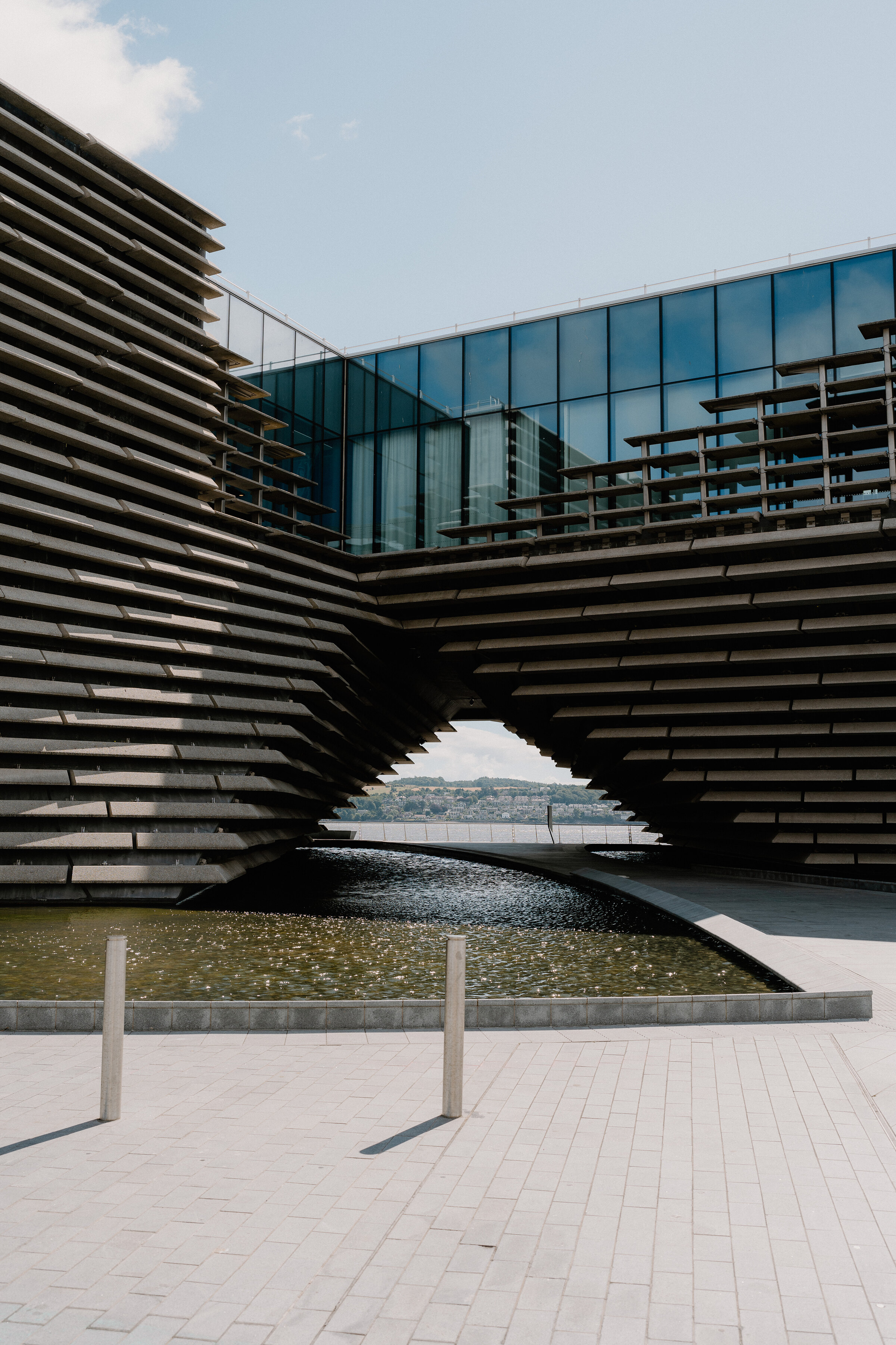A modern museum building's tiered concrete cladding curves overhead to frame a glass upper floor and a still water pool below.