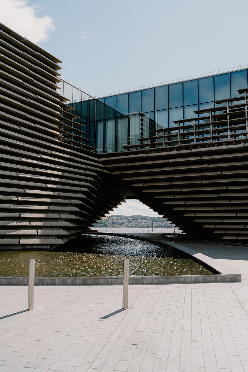 A modern museum building's tiered concrete cladding curves overhead to frame a glass upper floor and a still water pool below.