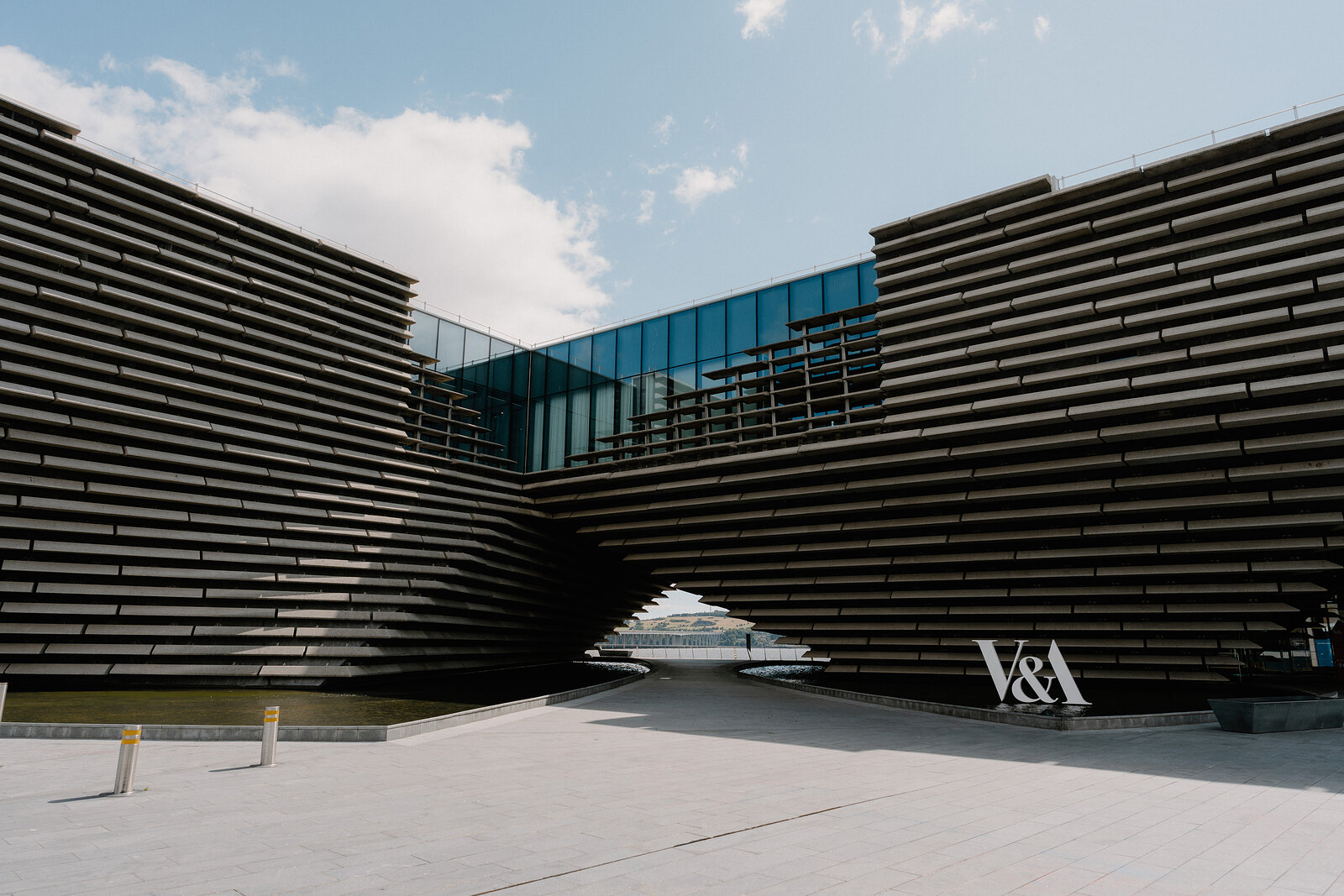 A modern museum building's tiered concrete cladding curves overhead to frame a glass upper floor, a sign reading 'V&A' mounted on the corner below.