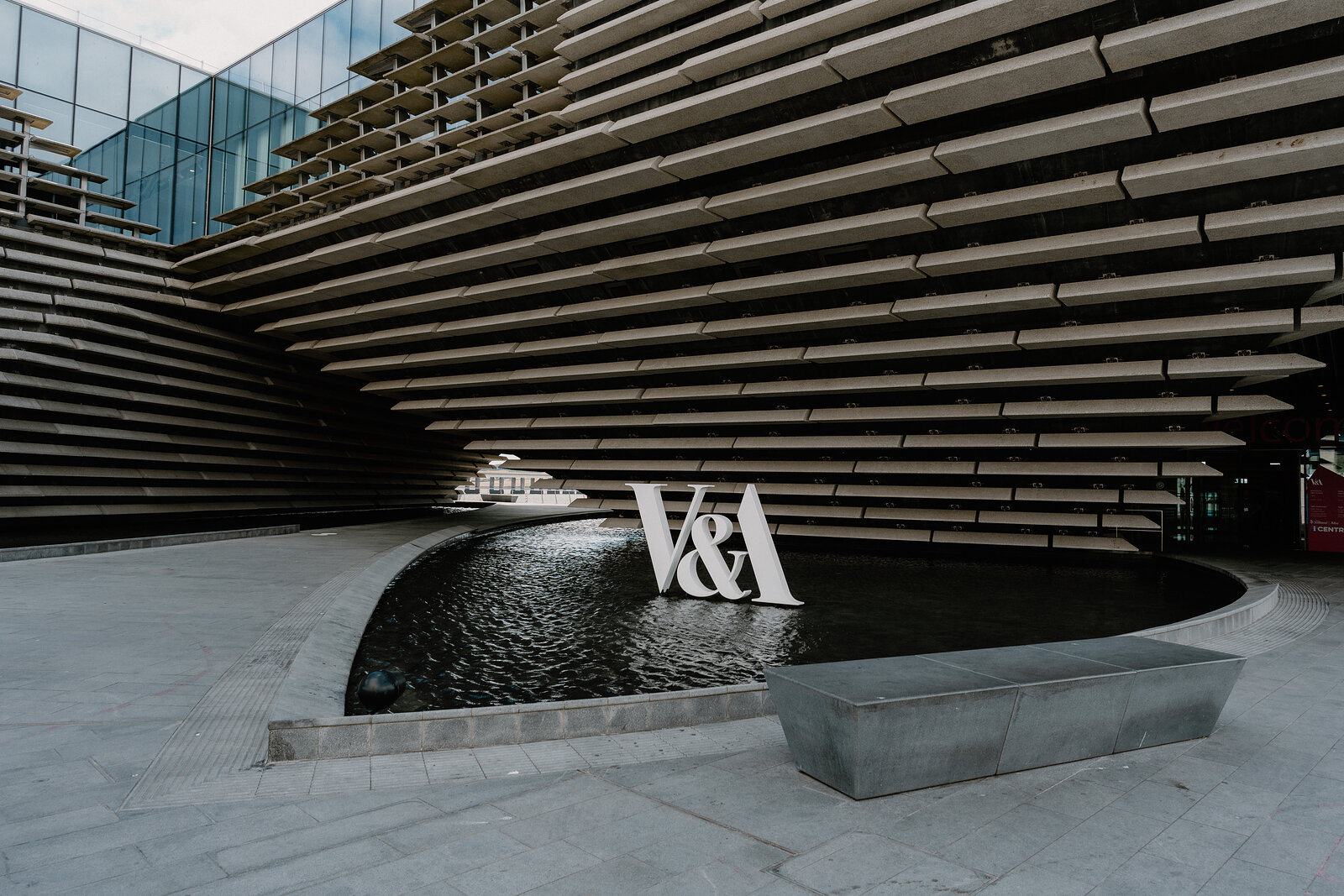 A large freestanding sign reading 'V&A' stands beside a still water pool beneath the tiered concrete overhang of a modern museum building.