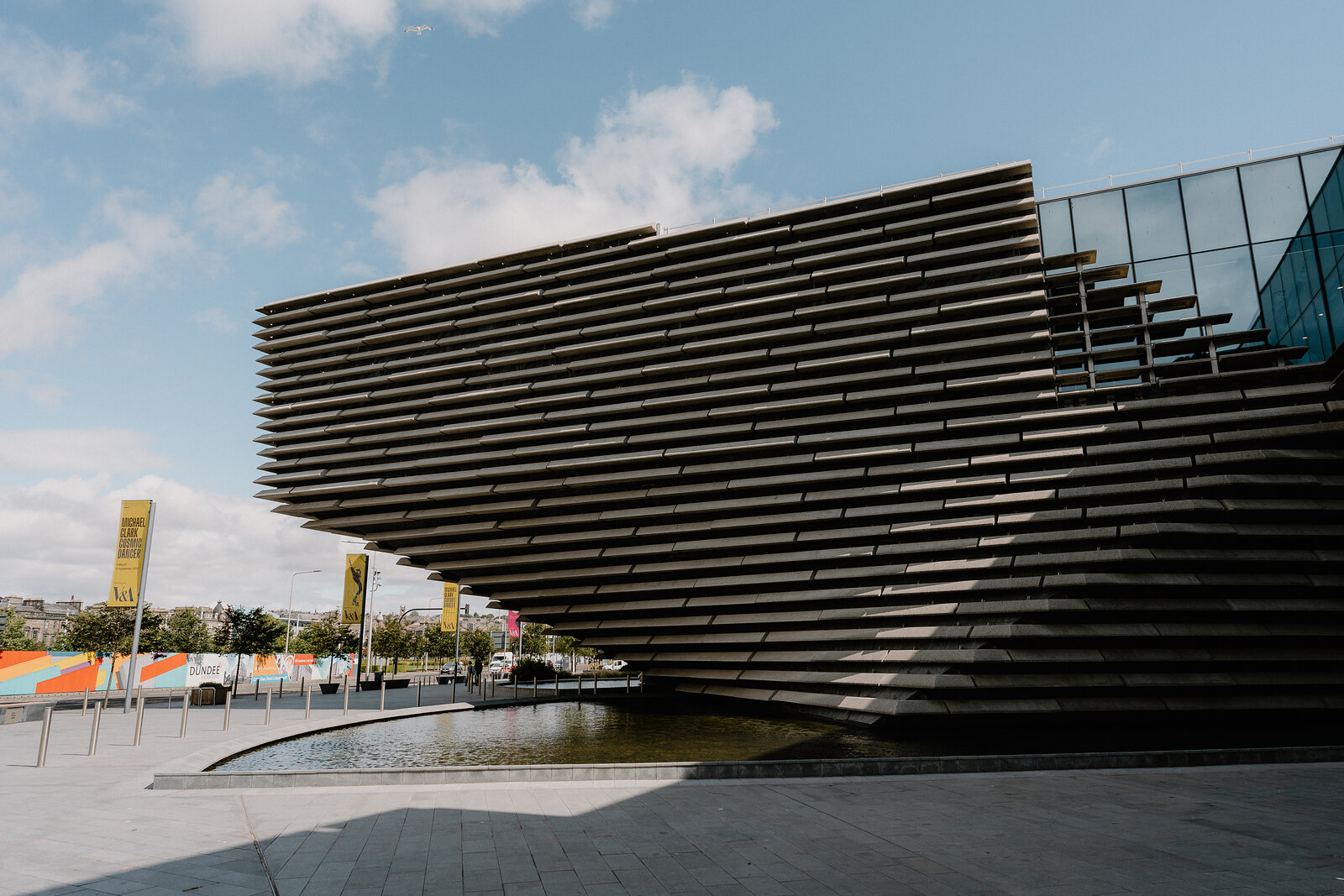 The tiered, ribbed concrete walls of a modern museum building curve above a paved plaza, banners and a water channel visible to one side.