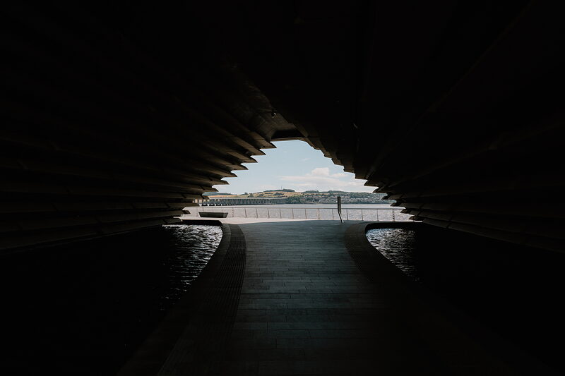 Looking through a dark tunnel of tiered concrete cladding toward a bright quayside with railings and a river beyond.