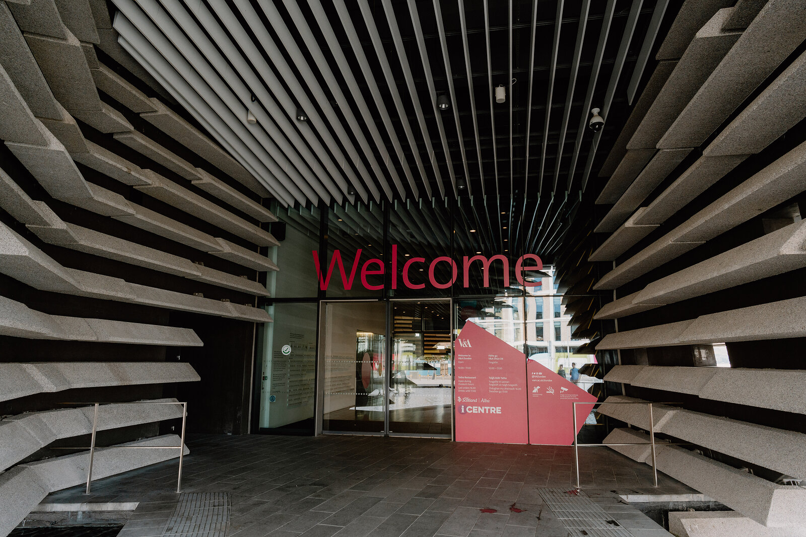 A red sign reading 'Welcome' hangs beneath the tiered concrete overhang at the entrance of a modern museum building.