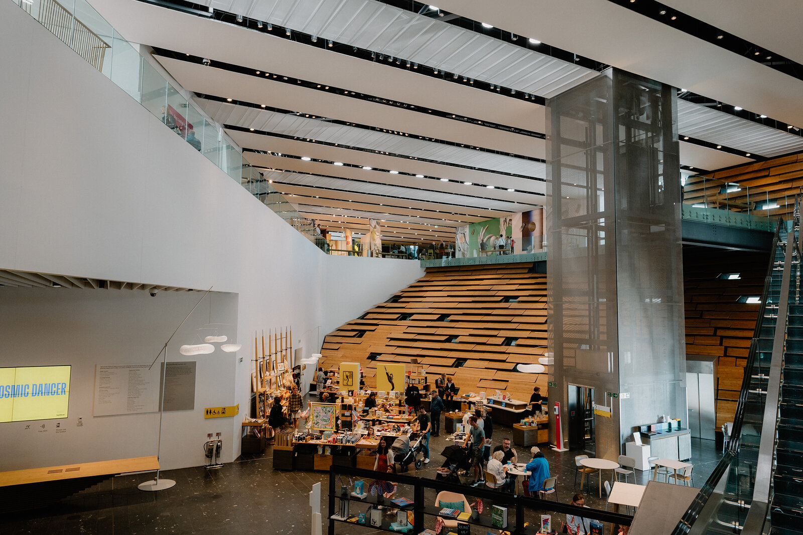 A double-height museum atrium with a timber-slatted mezzanine overlooks a gift shop and café below, a warning sign reading 'Cosmic Danger' mounted on a pillar.