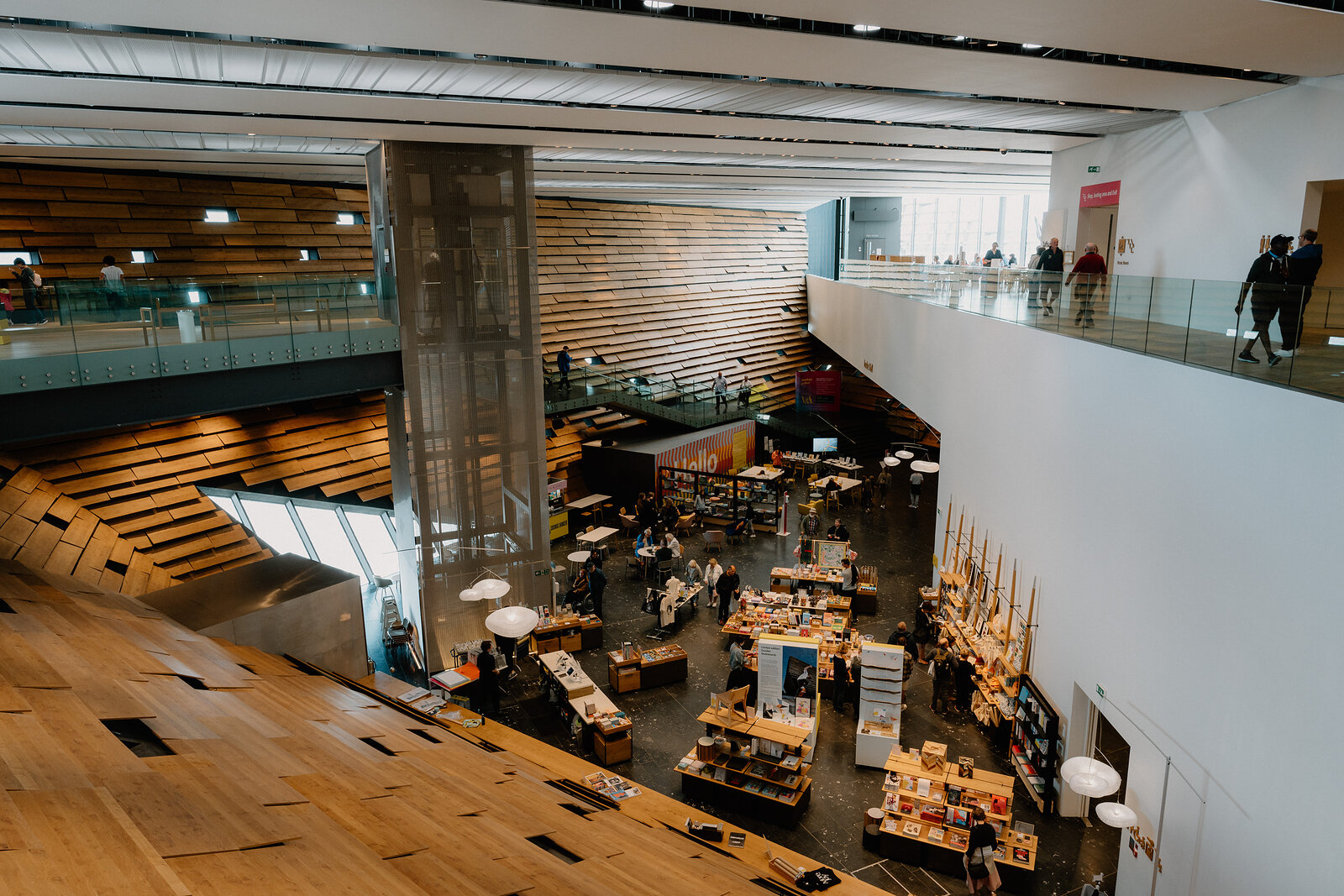 A double-height museum atrium with timber-slatted balconies overlooks a gift shop and café arranged with tables and shelving below.