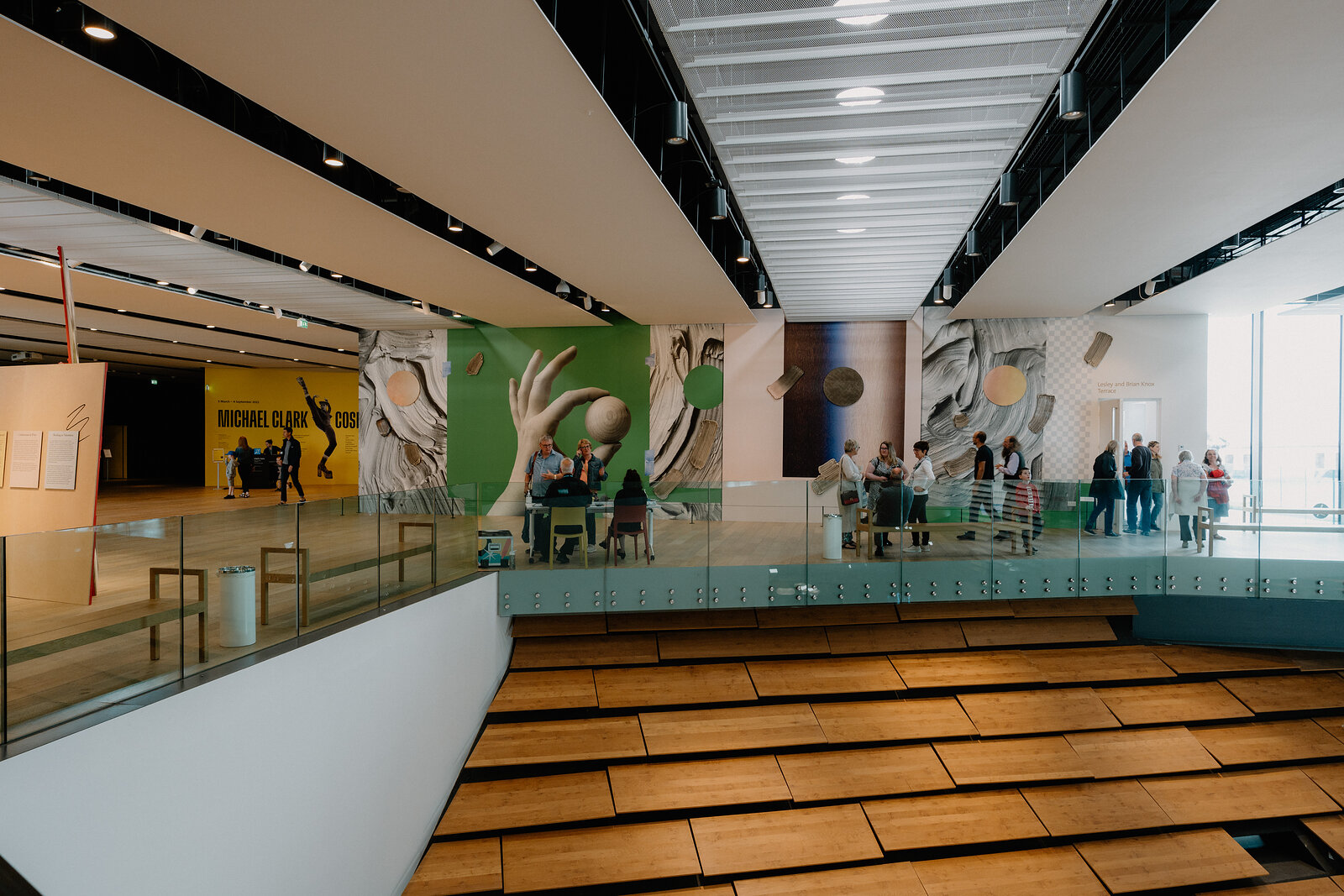 A museum atrium with wooden herringbone stairs looks across at illustrated wall murals of a diner scene and visitors browsing a gallery below.