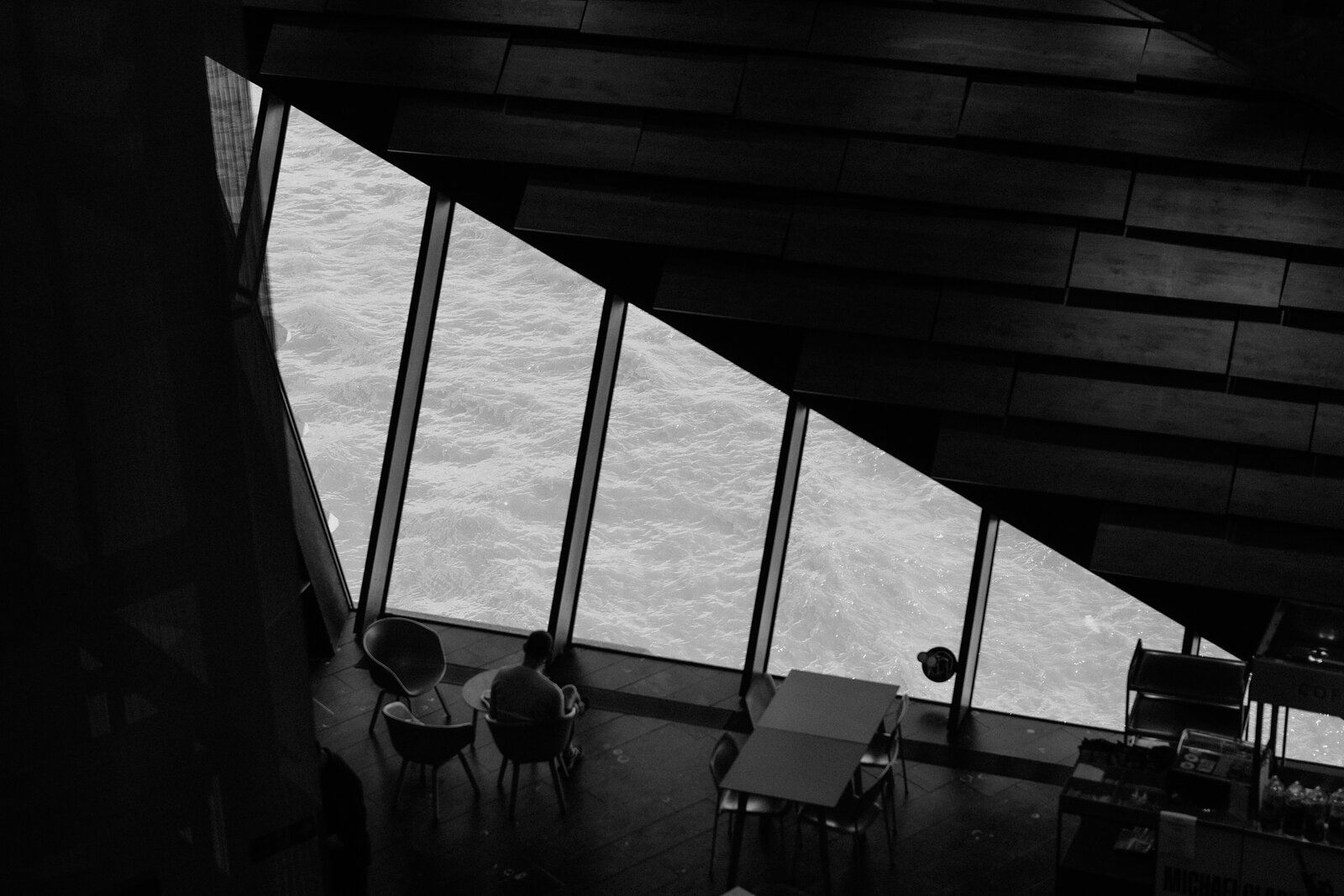 A monochrome photograph shows two people seated at a small table beside floor-to-ceiling windows overlooking a fast-flowing river, timber-slatted ceiling above them.