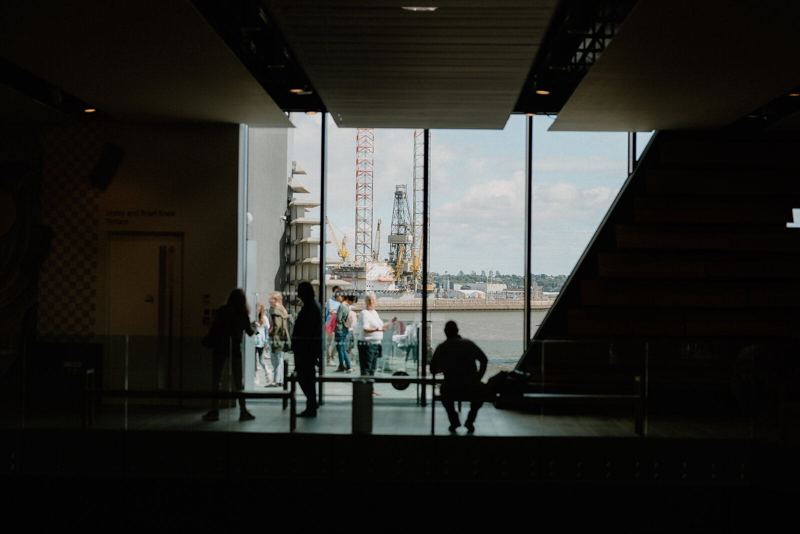 Visitors look out through a glazed archway toward moored ships on a river, a staircase and museum exhibit visible to one side.
