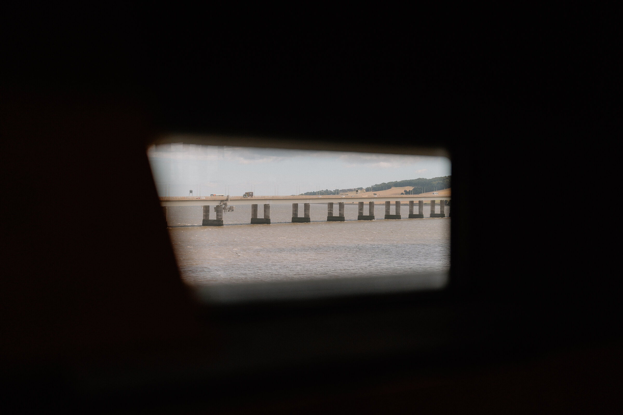 A rail bridge spanning wide water is framed through a small round window, the dark interior of a vehicle visible around the edges.