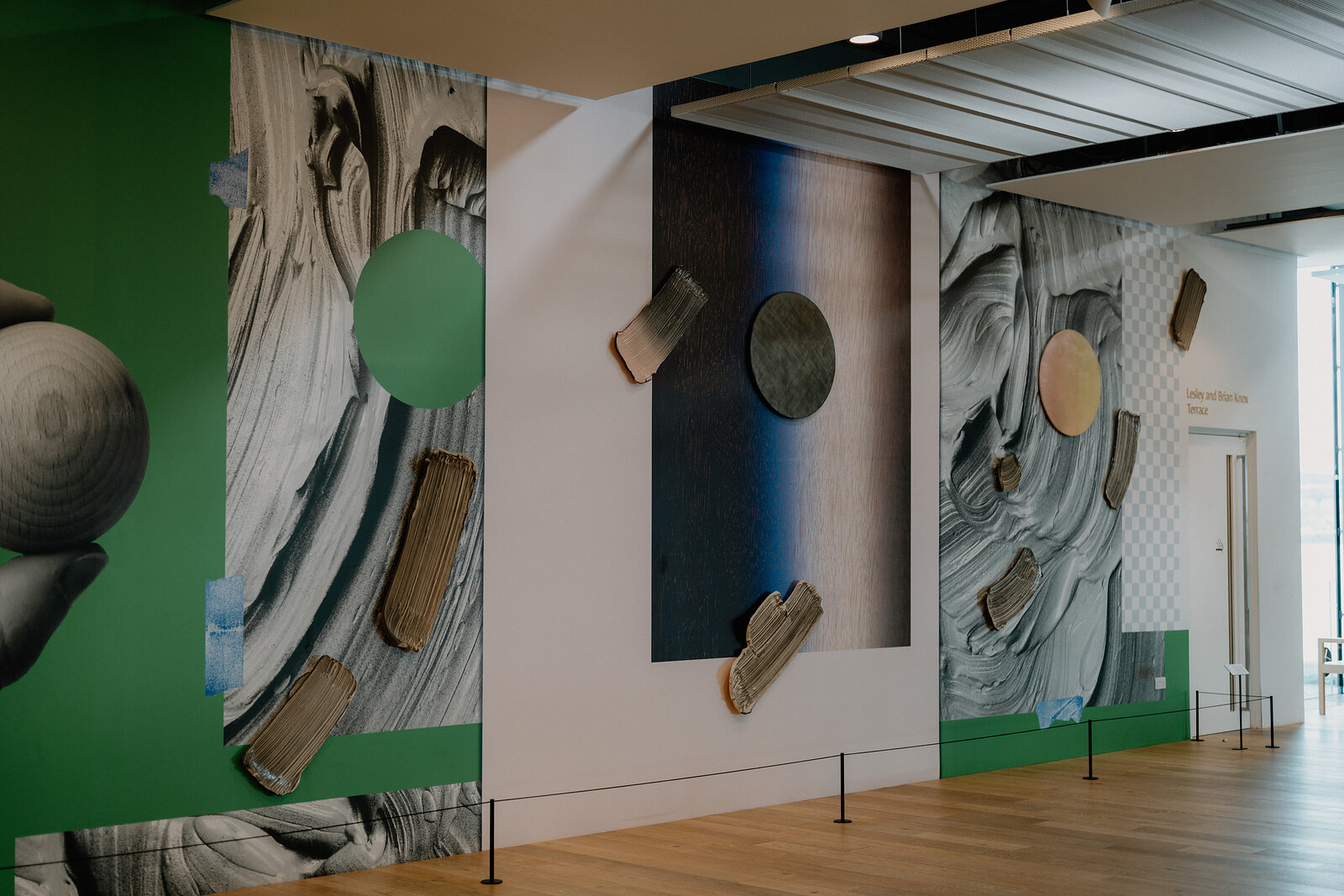 Large abstract photographic murals of driftwood and rock textures line a museum corridor beside a green wall and rope barriers.