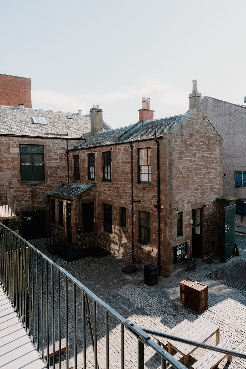 A sandstone tenement building with multiple chimney stacks overlooks a cobbled courtyard from a railed platform above.