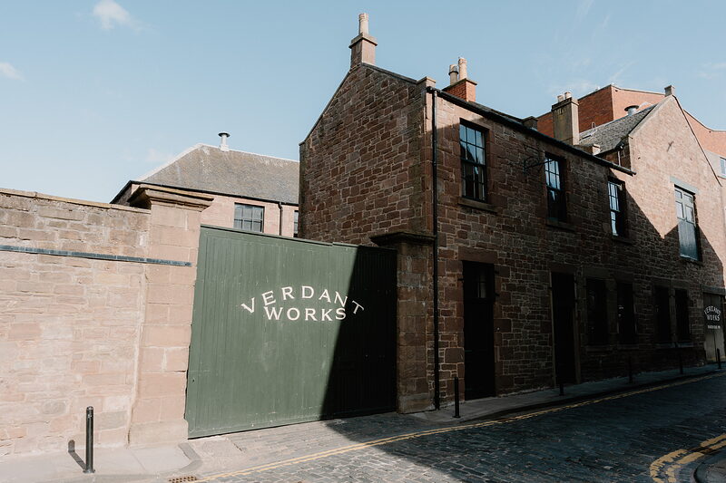 A sandstone building carries a sign reading 'Verdant Works' on a dark green gate, seen through overhanging tree branches beside a cobbled lane.