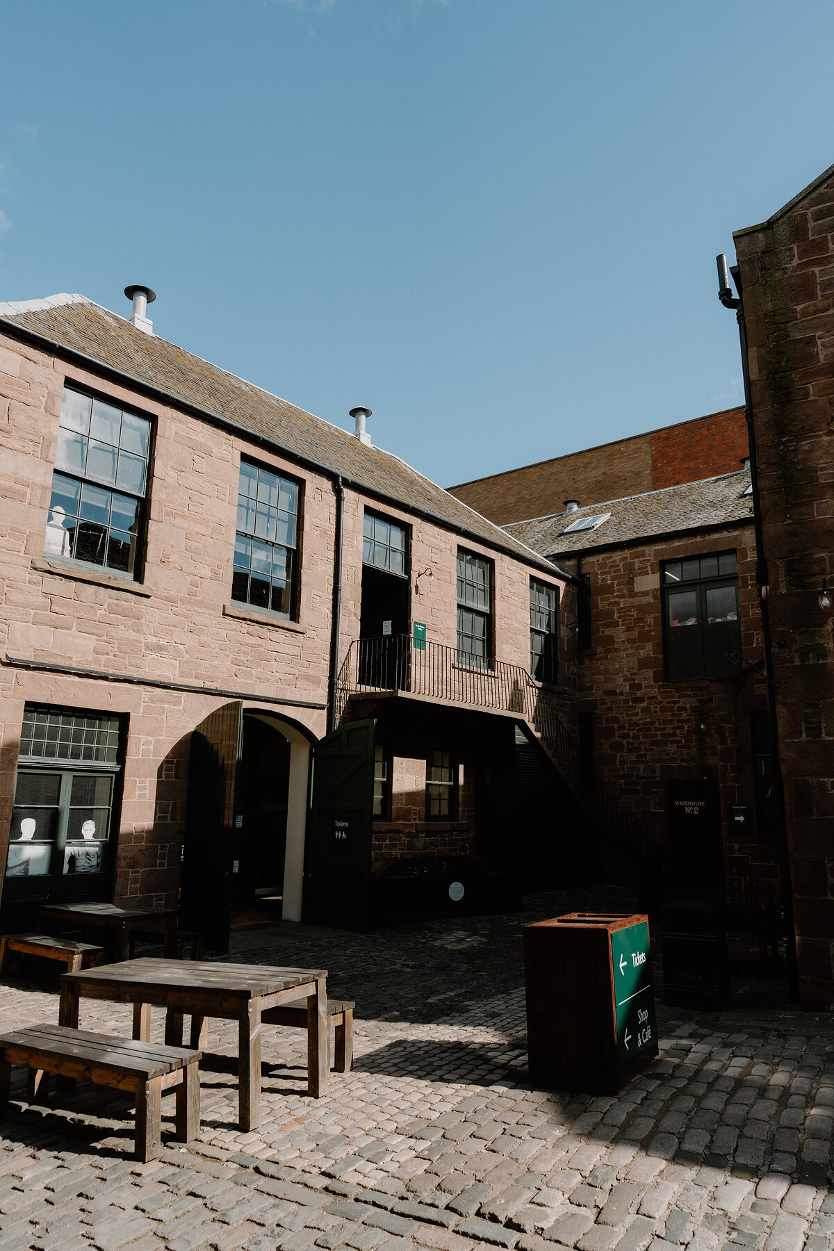 A sandstone courtyard building with an external iron staircase and arched doorways overlooks a cobbled yard set with wooden benches and a recycling bin.