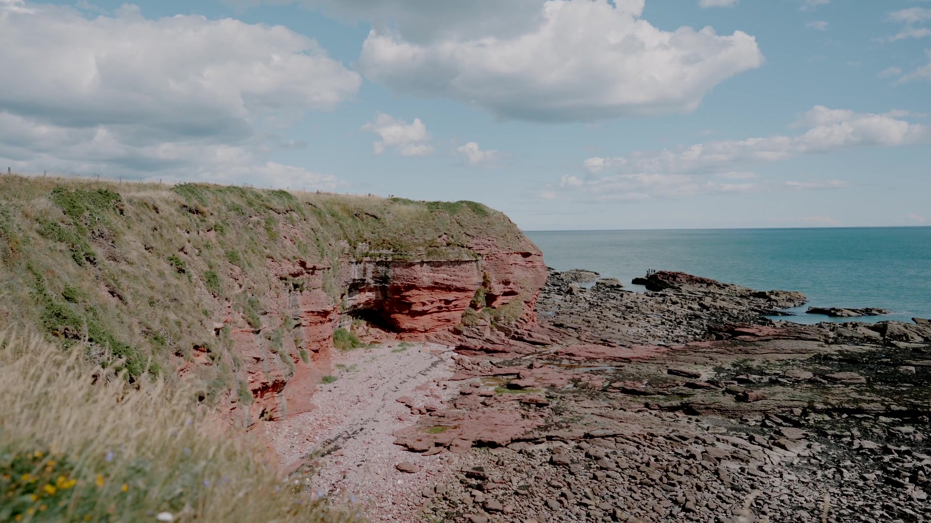 Red sandstone cliffs meet a rocky shore beneath a partly cloudy sky, calm turquoise sea stretching to the horizon.