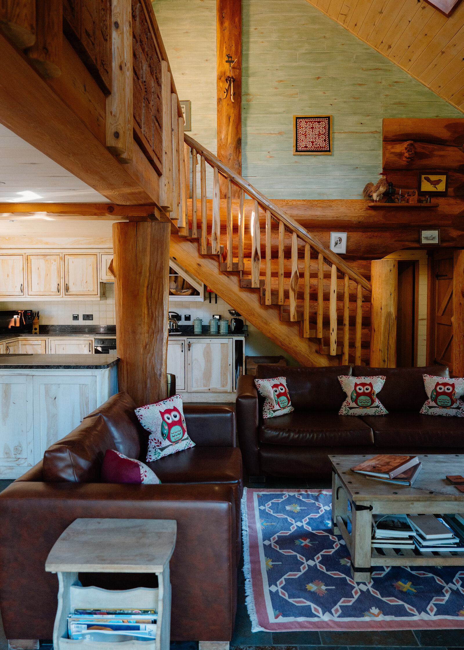 An open-plan log cabin interior with a wooden staircase and gallery rail rising past a stone-based log support, leather sofas scattered with owl-print cushions in front, and a fitted kitchen behind.