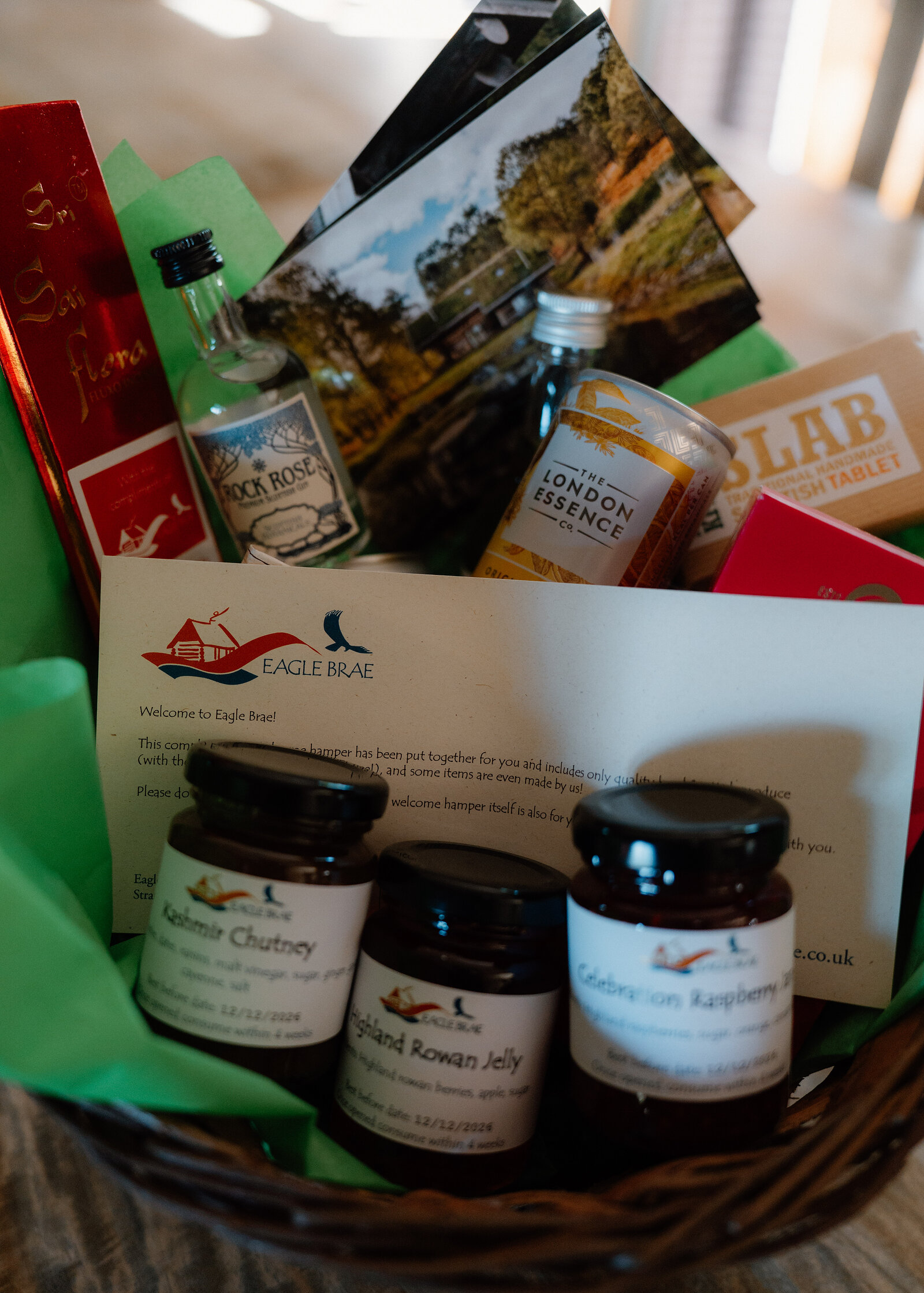 A wicker welcome hamper for Eagle Brae holding jars of Kashmiri chutney, rowan jelly and raspberry preserve, a miniature Rock Rose gin, a bottle of London Essence tonic, a bar of tablet, postcards and a printed welcome letter, set in green tissue paper.