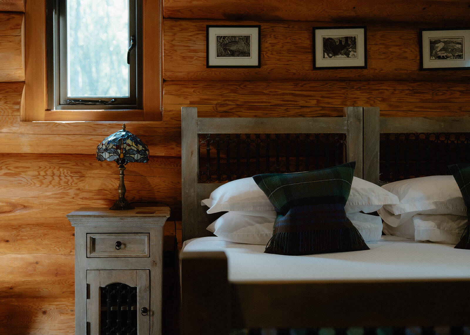 A tartan cushion sits among white pillows on a log-framed bed, a Tiffany-style lamp glowing on the bedside cabinet beside a window and three framed bird prints on the log wall above.