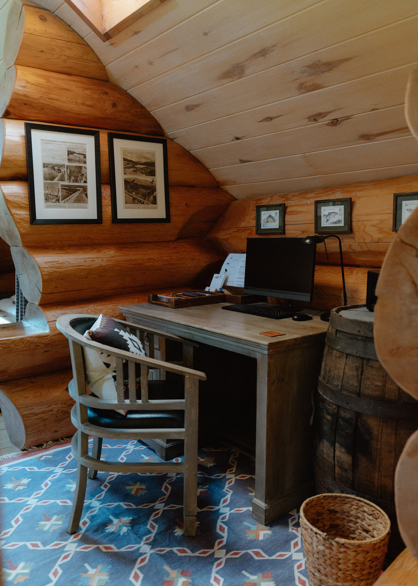 A sloped log-cabin nook holds a desk with a computer monitor beneath sepia photographs, a whisky barrel serving as a side table and a patterned rug underfoot.