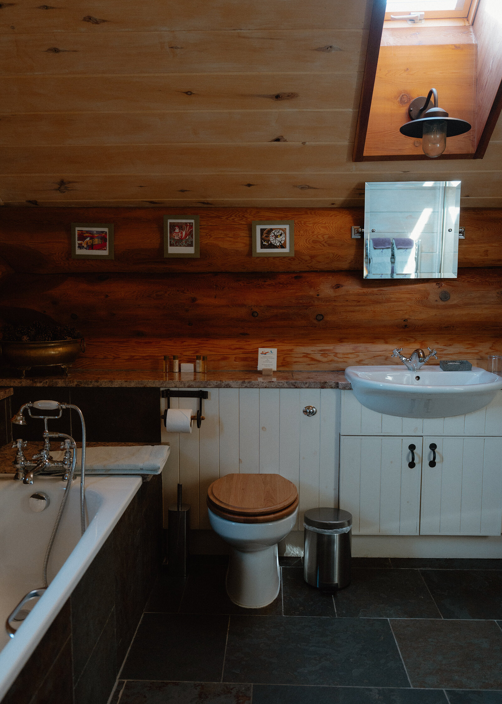 A roll-top bath with telephone-style taps sits beside a toilet with a wooden seat and a pedestal sink, framed botanical prints on the log wall and a skylight lighting the room above.