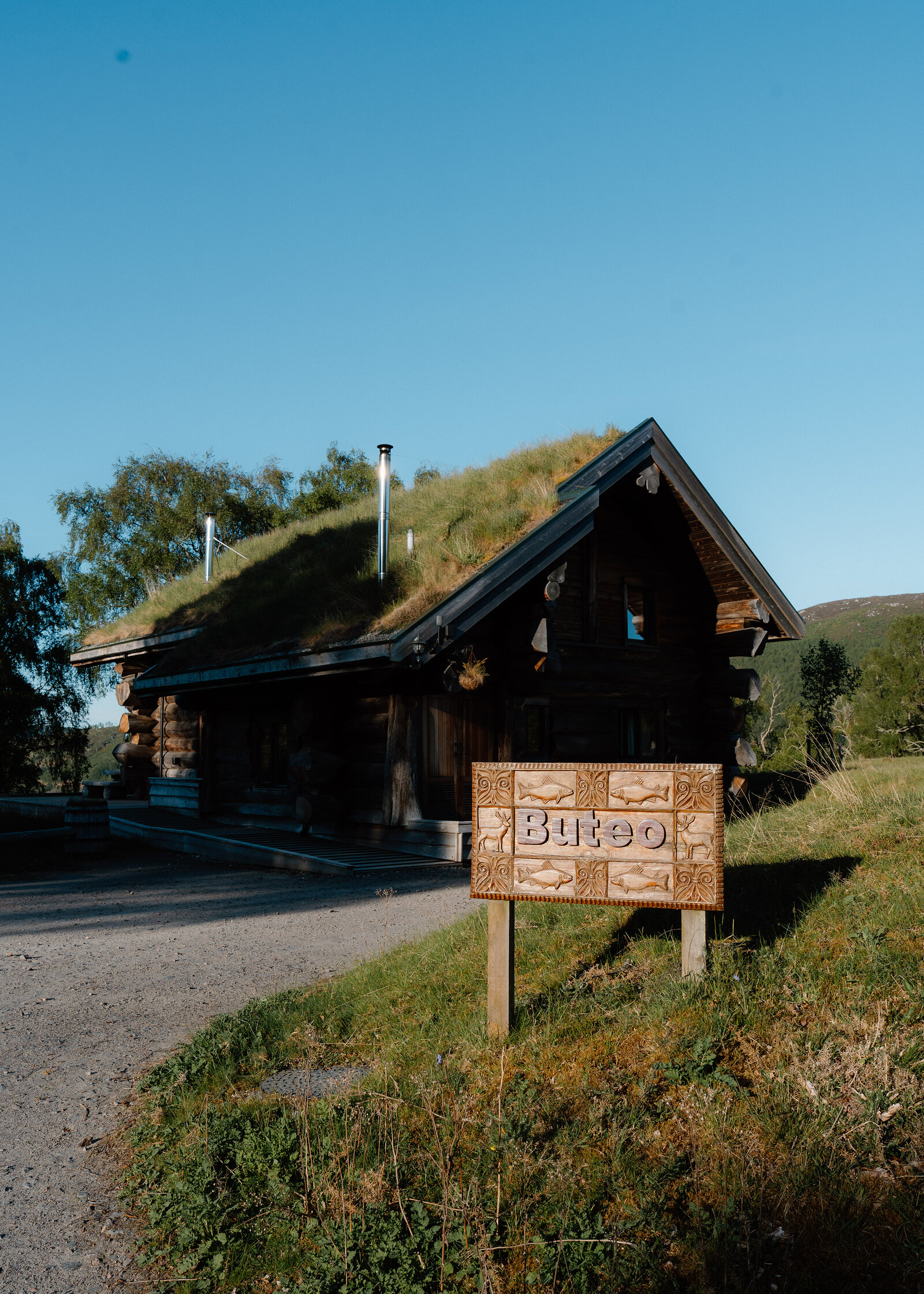A wooden sign reading Buteo stands on posts before a turf-roofed log lodge under a clear blue sky.