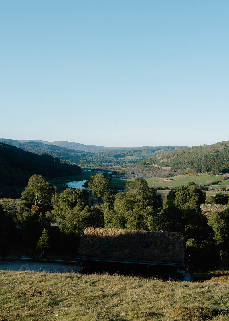 A river winds through a wooded valley towards distant hills, a stone building's roof visible among the trees in the foreground.