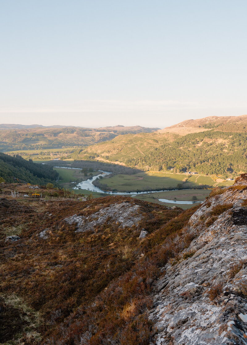 A river valley opens beyond a rocky, heather-covered foreground, forested slopes rising towards distant hills under a clear sky.