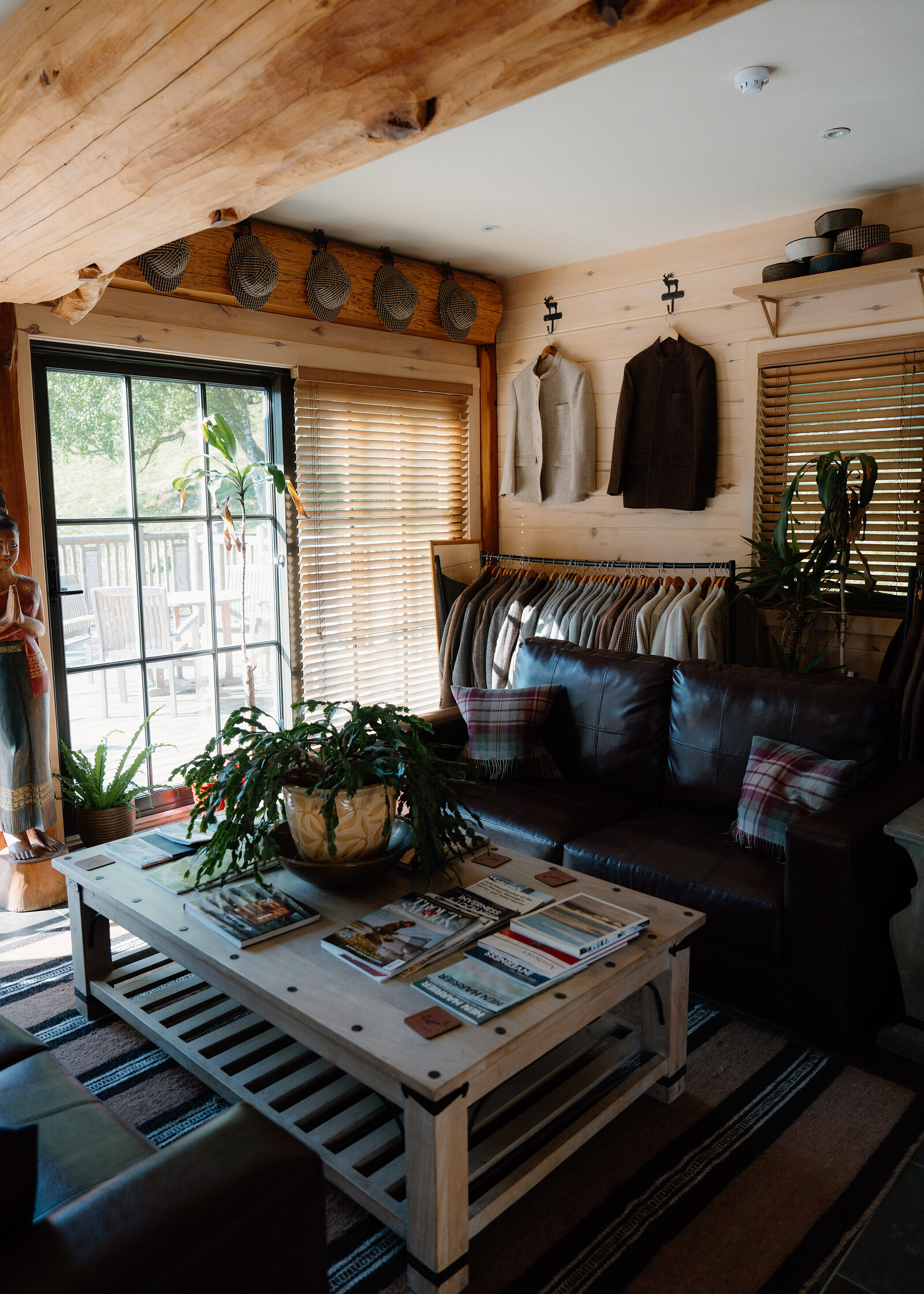 A rail of jackets and shirts hangs beside leather sofas and a low wooden coffee table stacked with magazines in a log cabin lounge, a wooden carved figure standing near the glazed door.