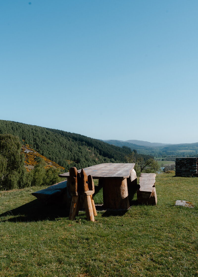 A carved wooden picnic table and benches stand on a hilltop meadow overlooking a forested glen and hills under a clear sky.