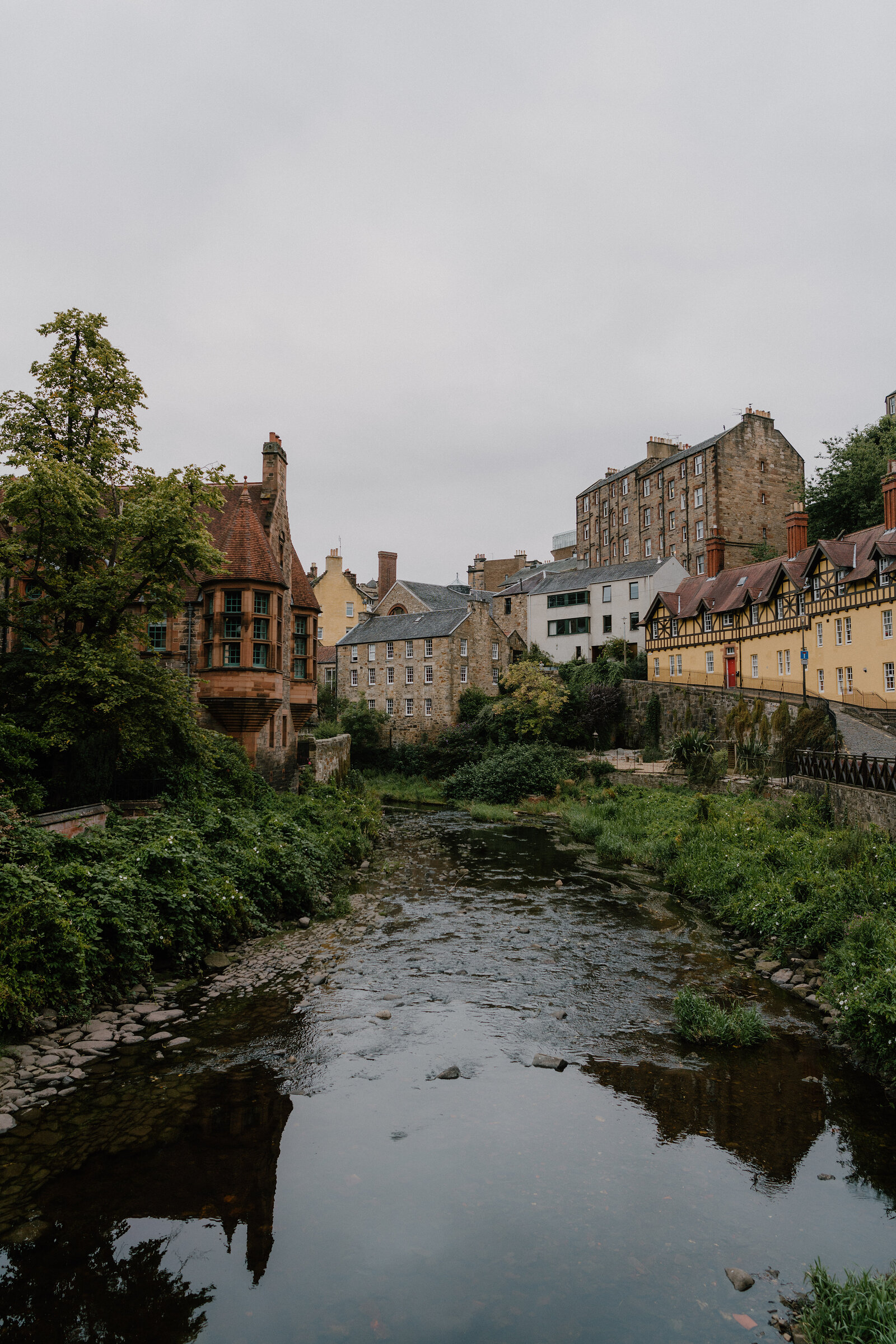 A row of colourful gabled buildings lines the Water of Leith, their reflections mirrored in the still water beneath overhanging trees.