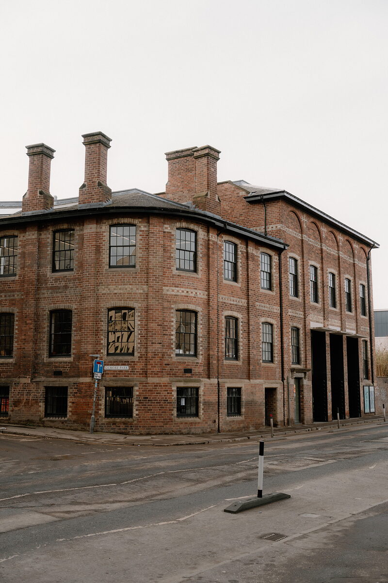 A large red brick Victorian warehouse with tall chimneys stands on a street corner, arched loading bays set into its ground floor.
