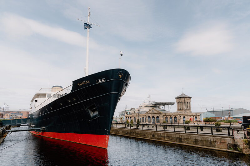 A black and red hulled ship named Fingal is moored at a dock, an ornate Victorian building and street lamp standing on the quay behind.