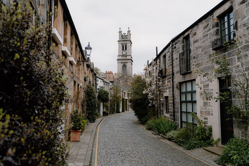 A cobbled mews lane lined with terraced stone houses and climbing plants, a church tower rising at the far end against a grey sky.