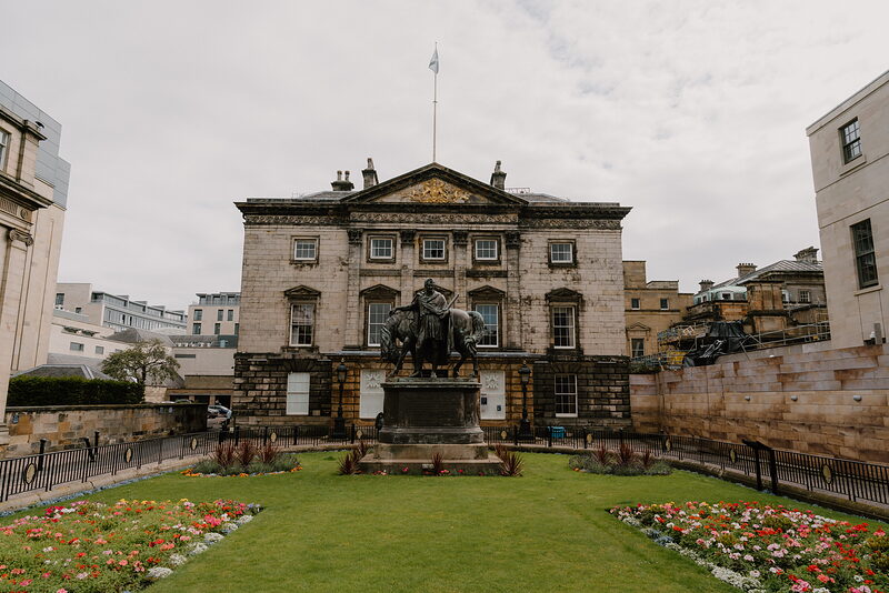 A pedimented sandstone building with a flagpole stands behind an equestrian statue on a plinth, flower beds and lawn in front.